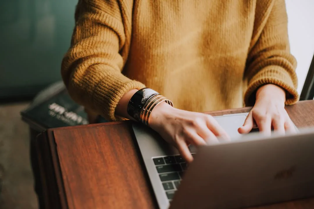 Faceless person in yellow sweater with bracelets sitting at a desk typing on a laptop